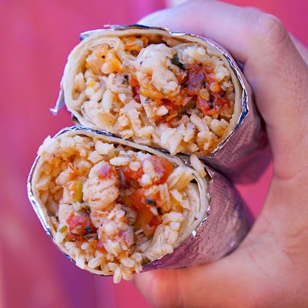 A hand holds a Baja Taqueria burrito, cut in half, revealing rice, shrimp, and salsa.