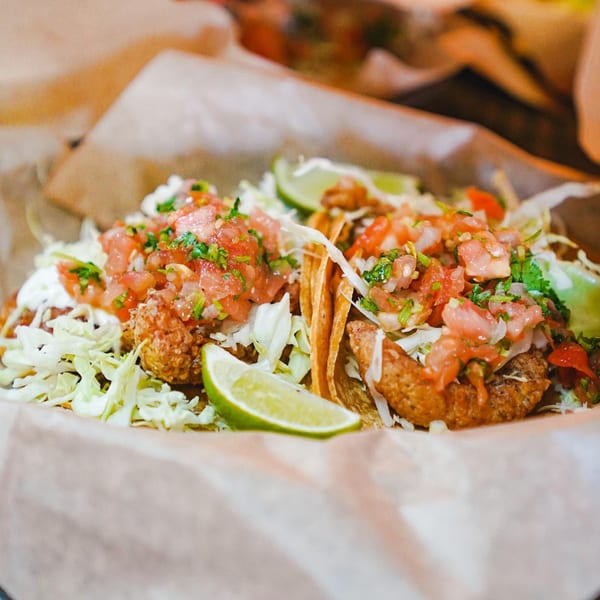 Two Baja Taqueria fish tacos with pico de gallo, shredded cabbage, and lime wedges.