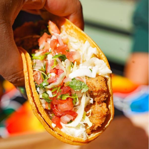 Hand holding a Baja Taqueria fish taco with crispy fish, cabbage, pico de gallo, and crema.