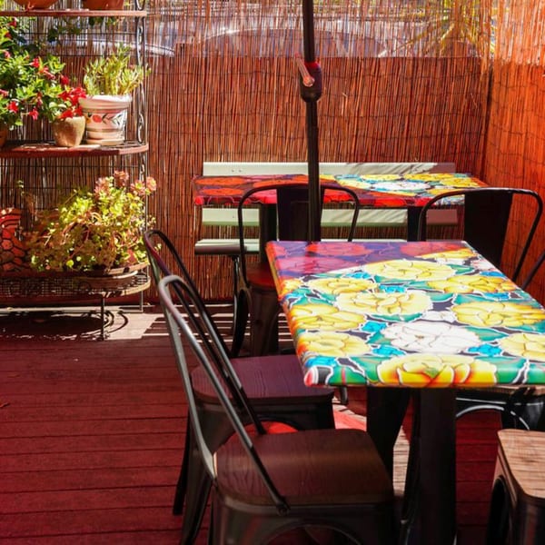 Outdoor patio with colorful floral tables, black chairs, potted plants, and a bamboo privacy screen.