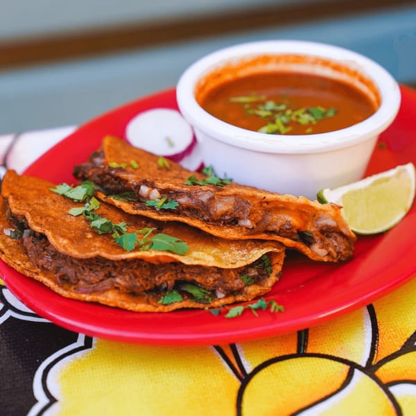 Three Baja Taqueria birria tacos with consome, lime, and radish on a red plate.